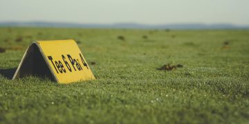 Close-up of a Tee 6 Par 4 sign on a golf course green field under clear skies.